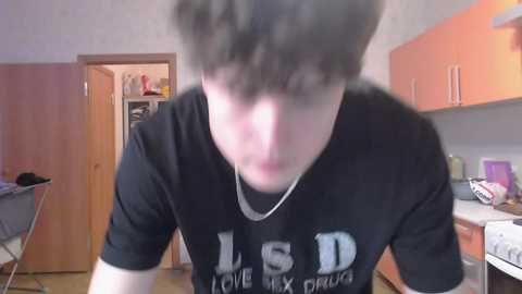 Media: Video of a young man with curly hair in a black T-shirt with white text, blurred, in a cluttered room with orange cabinets and a wooden door.