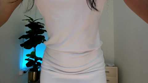 Media: A video of a woman's back in a white tank top, revealing a slim figure and black hair. Behind her, a potted plant and a dresser are visible.