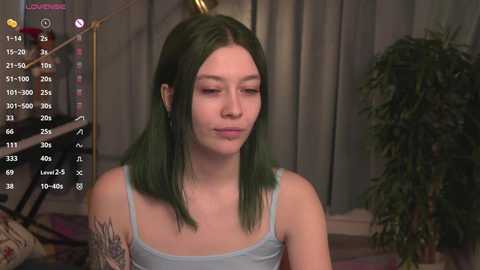 Media: A video of a young woman with fair skin and green hair, wearing a light blue tank top, standing indoors in a room with a potted plant and a bed.