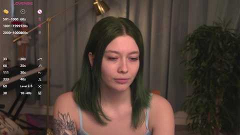 Media: Video of a young woman with fair skin and shoulder-length green hair, wearing a light blue tank top, sitting indoors. Background includes a potted plant and a gold lamp.