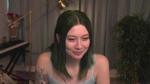 Media: Video of a young woman with fair skin and shoulder-length green hair, wearing a light blue tank top, smiling, in a cozy, dimly lit room with a potted plant and a desk in the background.