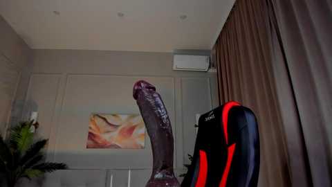 Media: Video of a large, realistic, dark purple dildo in a modern, beige-walled room with a black gaming chair, air conditioning unit, and abstract art on the wall.