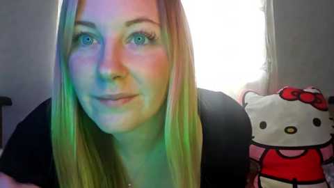 Media: A video of a young woman with straight, light brown hair, light skin, and green eyes, wearing a black top. She stands in front of a bright window and a Hello Kitty plush toy. The background is softly lit with a rainbow effect.