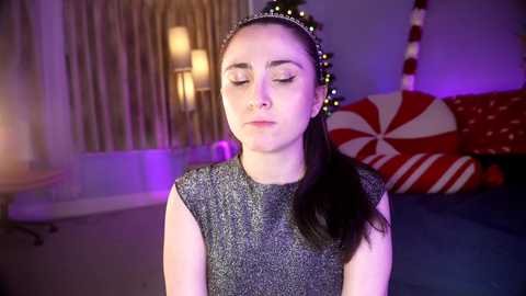 Media: Video of a young woman with light skin, dark hair, and closed eyes, wearing a glittery silver dress, seated indoors amidst festive decor with red and white candy cane pillows and glowing purple lights.