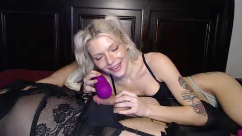 Media: Video of a smiling, blonde woman with tattoos, wearing black lingerie, using a purple sex toy on a woman with black lace lingerie, lying on a bed with dark wooden headboard.