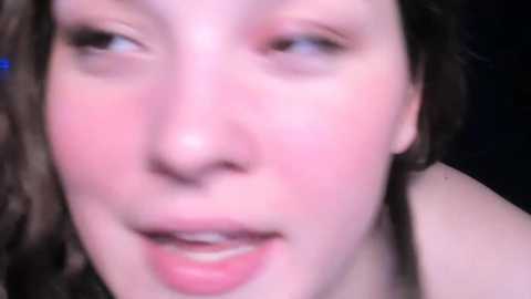 Media: A blurry close-up video of a fair-skinned woman with light brown hair and blue eyes, slightly smiling. She has a smooth complexion and is wearing minimal makeup. The background is out of focus, likely indoors with dim lighting.