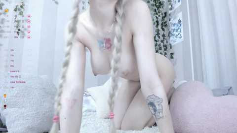 Media: Video of a pale-skinned woman with blonde twin braids, nude, with small breasts, tattoos, and a pink heart necklace, kneeling on a plush white rug, in a room with white curtains and fairy lights.