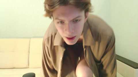 Media: A video of a young man with short, wavy brown hair, wearing a brown shirt, leaning forward on a cream-colored sofa. The background is a plain, light-colored wall.