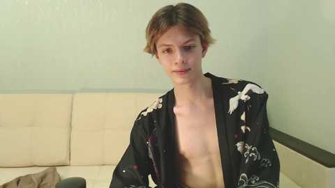 Media: Video of a young, fair-skinned, androgynous person with short, tousled brown hair, wearing a black robe with white patterns, sitting on a beige couch against a pale green wall, appearing contemplative.