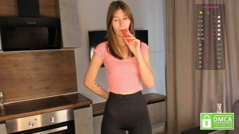 Media: A video of a slender, light-skinned woman with straight brown hair, wearing a pink crop top and black leggings, eating a red apple in a modern kitchen with wooden accents.