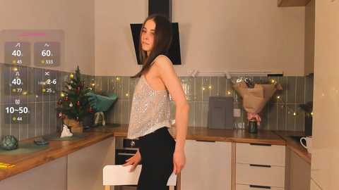 Media: A video shows a young woman in a kitchen, wearing a sleeveless, sequined top and black pants. She stands near a wooden countertop with a Christmas tree and wrapped presents.