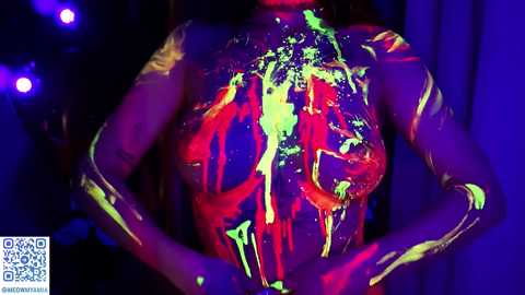 Media: Video of a person with dark skin, covered in vibrant, neon-colored body paint, standing against a dark backdrop. The person's hands are placed on their chest, highlighting the paint's vivid hues of red, green, and yellow.