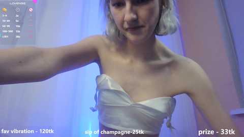 Media: Video of a pale-skinned, petite woman with shoulder-length, platinum blonde hair, wearing a white strapless dress, taken in a dimly lit room with blue and pink lighting. She appears to be in a video chat with a viewer, her arm extended.