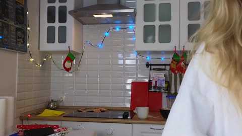 Media: A video of a modern kitchen with white subway tiles, red appliances, hanging lights, and festive Christmas decorations. A person with long blonde hair is seen from the back.