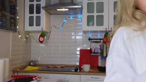 Media: Video of a modern kitchen with white cabinets, tiled backsplash, hanging fairy lights, red toaster, and a blonde woman in a white robe.