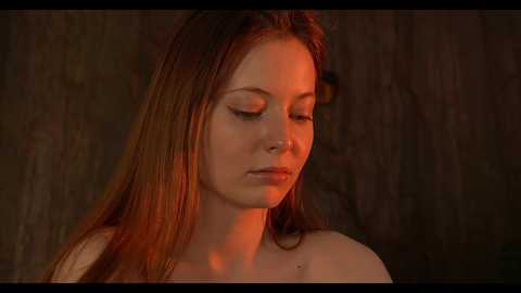 Media: A close-up video of a fair-skinned, red-haired young woman with closed eyes, bathed in warm, orange lighting, set against a rustic wooden backdrop.