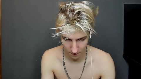 Media: Video of a pale-skinned, androgynous person with short, messy platinum blonde hair, wearing a silver chain necklace, listening to music with white earbuds. Background is a dark, muted gray wall.