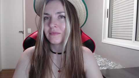 Media: A video of a young Caucasian woman with long, straight blonde hair, wearing a straw hat and a black choker, sitting on a red gaming chair in a minimalist room with white walls and a window.