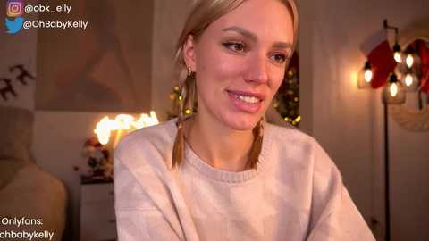 Media: Video of a smiling, fair-skinned woman with blonde hair in pigtails, wearing a beige sweater, indoors near a Christmas tree and string lights.