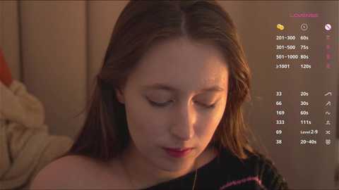 Video of a young Caucasian woman with long brown hair, wearing a black off-shoulder top, looking downward in a dimly lit room. Background includes a beige sofa and digital screens displaying statistics.