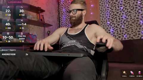 Media: Video of a muscular, bearded man with glasses, wearing a black tank top, playing a video game on a dark couch. Background features a leopard-print wall and a shelf with various items.