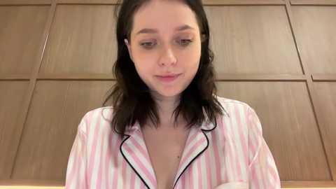 Media: Video of a young woman with fair skin, dark hair, wearing a pink and white striped pajama shirt, indoors with wooden paneled walls.