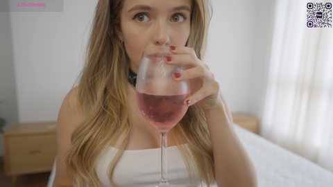 Media: Video of a young woman with long blonde hair, wearing a white tank top, drinking wine from a glass, in a bright, minimalist bedroom.