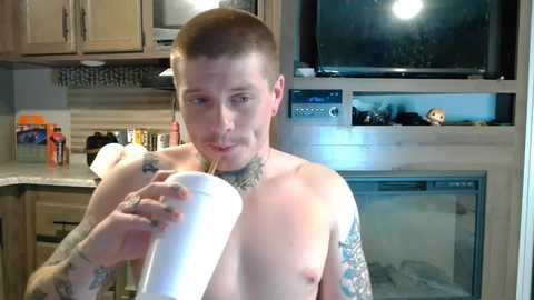 Media: Video of a shirtless, muscular, tattooed man with short hair, drinking from a white cup, in a modern kitchen with beige cabinets, microwave, and a small stuffed animal on the counter.