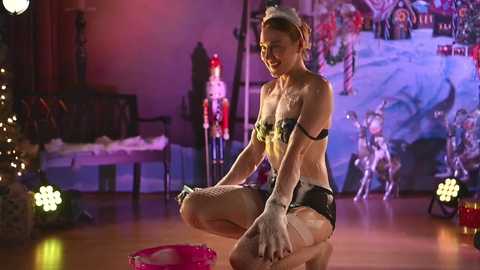 Media: Video of a blonde woman in a revealing black and white maid outfit with white gloves, squatting on a wooden floor, surrounded by festive Christmas decorations and metallic reindeer statues.