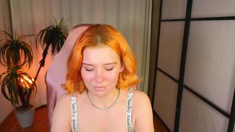 Media: Video of a fair-skinned young woman with shoulder-length, vibrant orange hair, wearing a white tank top with black patterns, standing in a softly lit room with a potted plant and shoji screen.