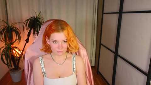 Media: Video of a young woman with vibrant orange hair, wearing a white tank top with silver trim, seated indoors. Background features a lighted potted plant, beige curtains, and a shoji screen.