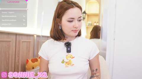 Media: Video of a young woman with shoulder-length auburn hair, wearing a white t-shirt with a cartoon dog graphic, and silver hoop earrings. She stands in a cozy, brightly-lit room with white walls and wooden paneling.