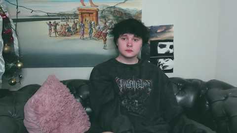 Media: Video of a young person with dark curly hair, wearing a black Vampire Weekend t-shirt, sitting on a dark leather couch. The background features a colorful mural and string lights.