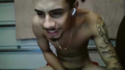 Media: Video of a shirtless, tattooed, light-skinned man with medium-length, curly hair, wearing white earbuds, leaning forward, with a light-colored background featuring a beige towel and a garage door.