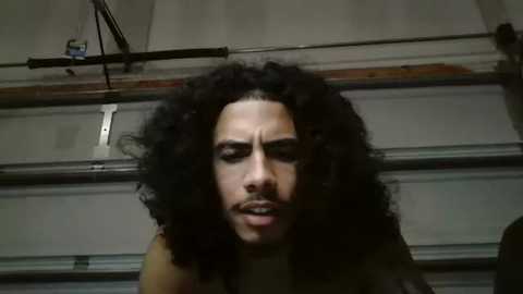 Media: A video of a man with voluminous curly black hair and a beard, looking distressed in a dimly lit garage with a metal door and electrical wiring visible.