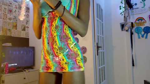 Media: Video of a person wearing a bright, tie-dye mini dress in a colorful, psychedelic pattern. The background shows a cluttered room with posters, a TV, and a door.