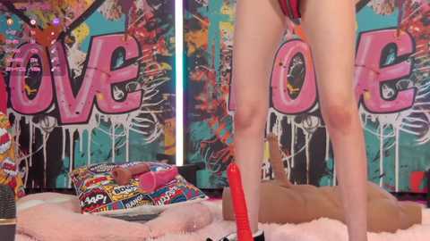 Media: Video of a person standing in a room with graffiti walls, holding a red dildo. The room is cluttered with colorful pillows and a pink rug.