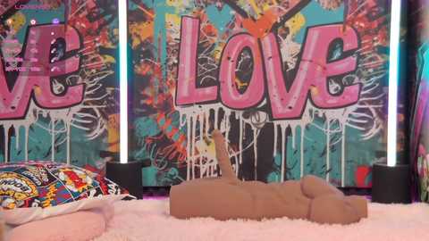 Media: Video of a nude mannequin lying on a pink shag rug, with a colorful umbrella in the background, surrounded by graffiti-covered walls with the word \"Love\" prominently displayed.