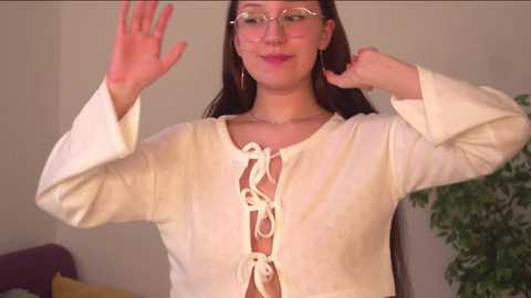 Media: Video of a smiling woman with fair skin, long dark hair, wearing glasses and a white, lace-up top, raising her arms in a casual indoor setting.