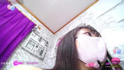 Media: Video of a young woman with long black hair, wearing a pink face mask, standing in a room with white marble walls, a purple curtain, and a framed artwork.