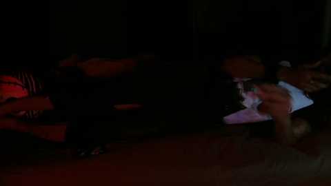 Media: Video of a dimly lit, nighttime outdoor scene featuring two people, one in a red shirt, lying prone on the ground. The other, in a white shirt, is kneeling, possibly tending to the prone person.