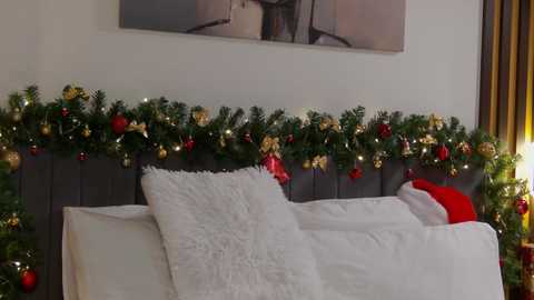 Media: Video of a cozy bedroom with a white bed, gray headboard, festive green garland adorned with red, gold, and silver ornaments, and a plush white pillow.