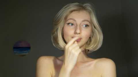 Media: Video of a fair-skinned, blonde woman with shoulder-length hair, wearing a contemplative expression, touching her lips. She is topless, with a blurred, dark background.