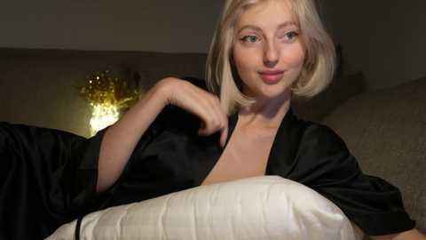 Media: Video of a fair-skinned woman with short blonde hair, wearing a black satin robe, resting on a white quilted pillow, in a dimly lit, cozy bedroom setting.