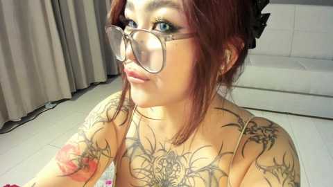 Media: Video of a fair-skinned woman with red hair, wearing glasses and large tattoos on her chest and arms, sitting in a modern, white room with gray curtains and a white couch.