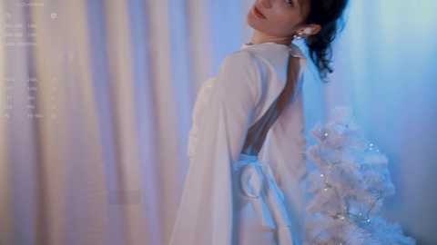 Media: Video of a woman in a flowing white dress with a sheer back, against a soft, pastel-colored backdrop featuring sheer curtains and a sparkling white tree. The lighting is soft, creating a serene, ethereal atmosphere.