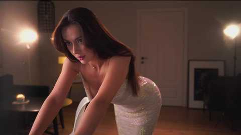 Media: Video of a brunette woman in a tight, glittering white dress, leaning forward in a dimly lit, modern living room with wooden floors and warm lighting.