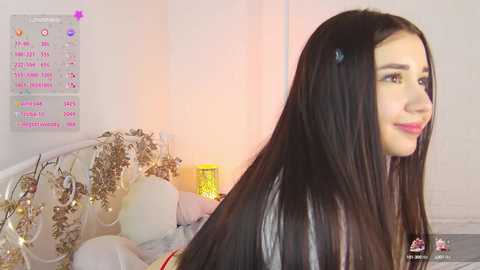 Media: A video of a young woman with long, straight black hair and light skin, smiling in a cozy, pink-walled bedroom with a bed adorned with white and gold decorations and fairy lights.