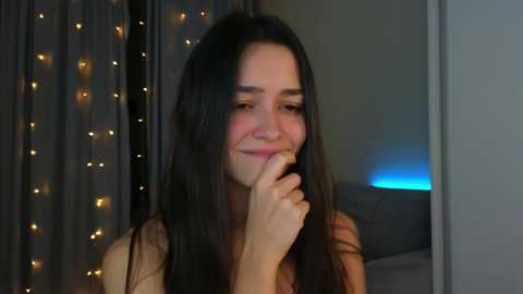 Media: Video of a young woman with long black hair, fair skin, and a small smile, touching her finger to her lips. Background features a curtain with fairy lights and a glowing blue light source.