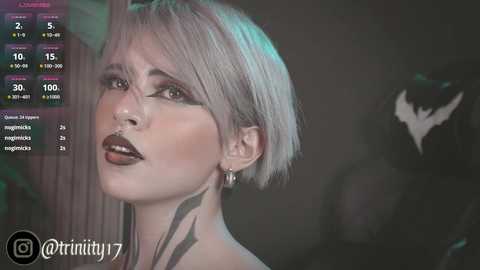 Media: Video of a woman with short, platinum blonde hair, heavy makeup, and a black tattoo on her neck, wearing a black top. Background features a dark, shadowy room with a Batman logo.